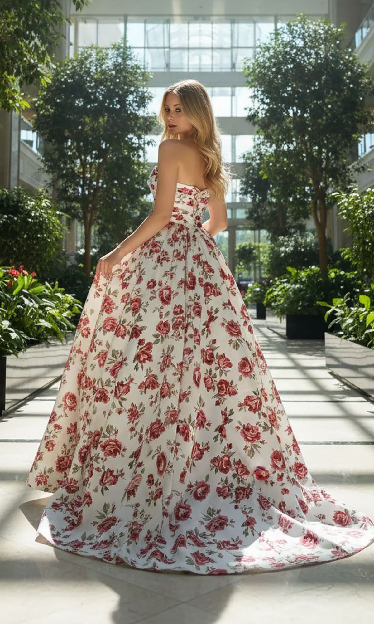 A woman wearing the Amelia Couture 8070 long prom dress, featuring a strapless red floral design, stands indoors among lush green plants with natural light streaming through tall windows.
